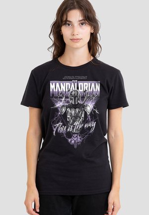 Young woman wearing a black T-shirt with The Mandalorian character and text "This is the way" on a plain background.