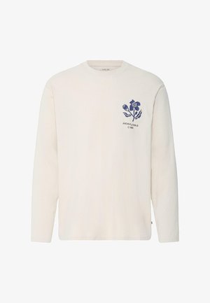 Roomkleurig shirt met lange mouwen en ronde hals, met een kleine blauwe bloemafbeelding en de tekst "ARCHIVE of SOLID © 1999" op de linkerbovenkant van de borst.