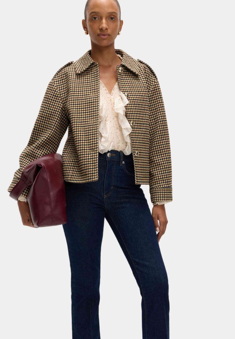 Bruine en beige ruitjesjas met een kraag, voor knopen, gecombineerd met een ruffle witte blouse en donkere spijkerbroek. Houdt een bordeauxkleurige tas vast.