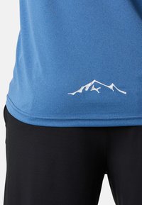 Blå sportskjorta med en mjuk textur, försedd med en vit bergslogotyp vid nederkanten, tillsammans med svarta byxor.