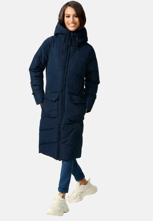 Navy puffer coat with a long design, hood, and front zip. Features two large front pockets and a quilted texture. Paired with jeans and sneakers.
