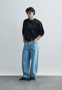 Čierny sveter s vyrazeným logom, svetlomodré široké rifle a čierne tenisky. Model stojí s rukami v vreckách na jednoduchom pozadí.