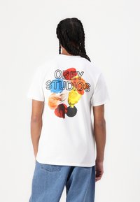 White cotton t-shirt featuring "OBEY STUDIOS" in black text on the back with colorful paint-like splotches in red, blue, yellow, and black.