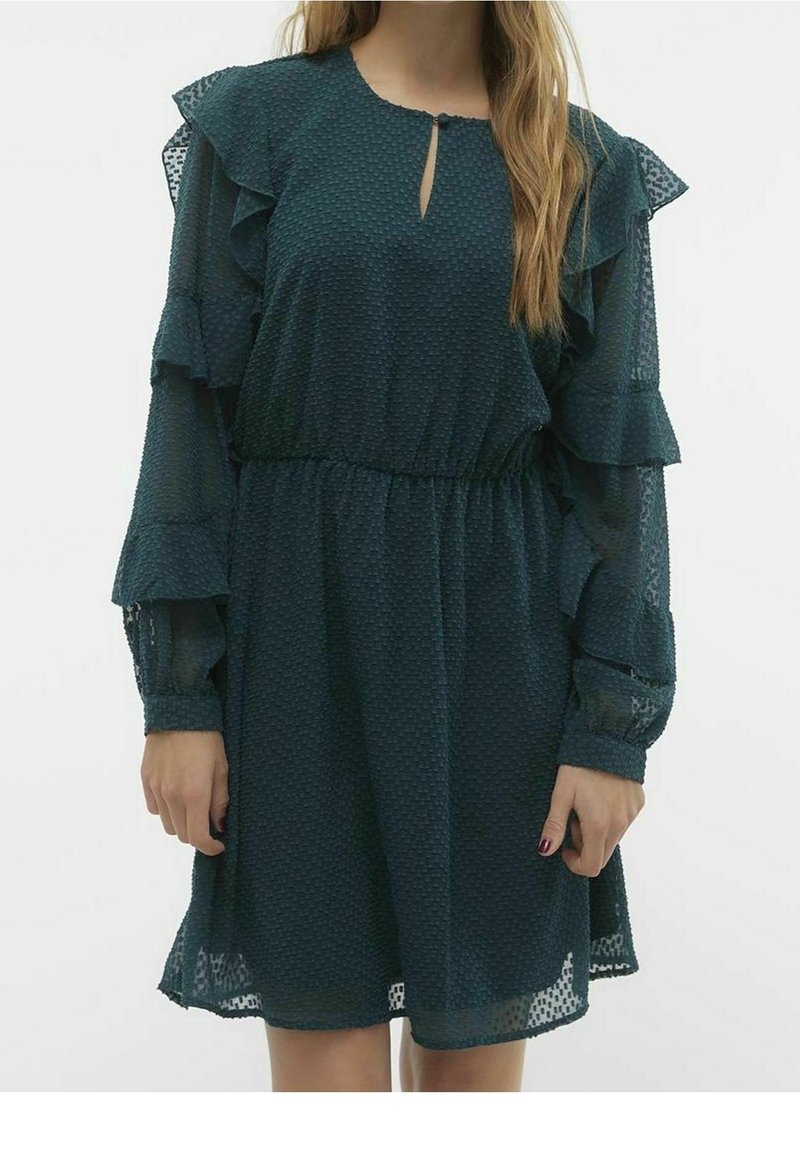 Femme portant une robe bleu foncé texturée avec de longues manches à volants et un petit décolleté en forme de trou de serrure à l'avant.