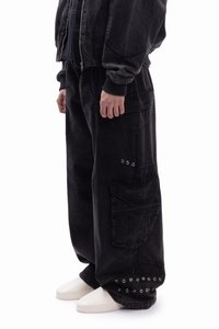 Črne kavbojke iz trapera s širokimi stranskimi žepi, z zadrgami na pritrdilih in kovinskimi okraski na spodnjem robu. Sproščen kroj s teksturirano strukturo.