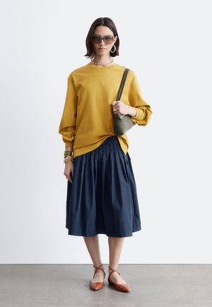 Kobieta w okularach przeciwsłonecznych, żółtym musztardowym topie z długim rękawem, granatowej spódnicy midi, brązowych płaskich butach, złotej biżuterii i trzymająca szarą torebkę na ramię.