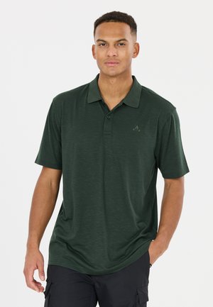 Mann trägt ein dunkelgrünes Polohemd mit kurzen Ärmeln und dezentem Logo, steht mit einer Hand in der Tasche einer schwarzen Cargohose vor einem weißen Hintergrund.