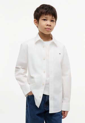 Jongen met kort donker haar, die een wit overhemd draagt over een wit T-shirt en blauwe spijkerbroek, staand met één hand in de zak.