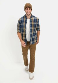 Geruite shirt in blauw en beige tinten, wit T-shirt, bruine corduroy broek en lichte sneakers; draagt een bruine gebreide beanie.