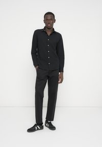 Camisa negra abotonada con mangas largas, pantalones negros y zapatillas negras con rayas blancas. Tejido liso, corte ajustado y diseño minimalista.
