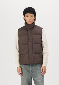 Brun polstret vest med høj krave og lodret syning, båret over en cremet sweatshirt. Modellen står foran en ensfarvet hvid baggrund.