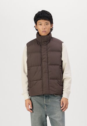 Gilet rembourré marron avec col montant et coutures verticales, porté sur un sweat-shirt crème. Le modèle se tient devant un fond blanc uni.