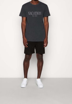 Muž oblečený v černém tričku "Vacation Club Est. 2016", černých šortkách a bílých teniskách, stojící proti jednoduchému světlému pozadí.