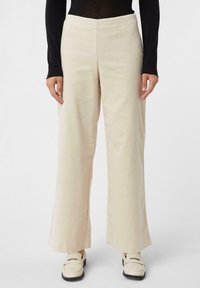 Spodnie o szerokich nogawkach w jasnym beżowym kolorze, z teksturowanej tkaniny i gładkim pasem. Stylizowane z dopasowanym czarnym topem z długim rękawem oraz białymi butami.