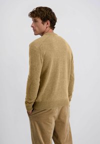 Gebreide tan kleurige trui met een ronde halslijn en een geribde zoom; gecombineerd met beige corduroy broeken, met handen in de zakken, bekeken van achteren.