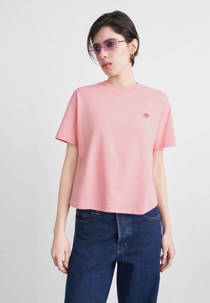 Vrouw draagt een loszittend roze T-shirt met een klein logo en donkerblauwe jeans, geaccentueerd met paarsgetinte bril en gouden hoepeloorbellen.
