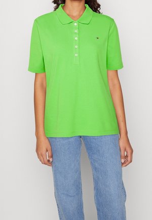 Personne portant un polo vert vif à manches courtes avec des boutons blancs et un jean bleu clair sur un fond uni.