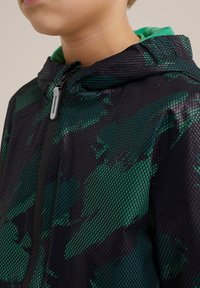 Černá a zelená maskáčová bunda na zip s síťovanou texturou, kapucí a kontrastním zeleným podšíváním. Obsahuje zipový přední díl a prodyšnou látku.