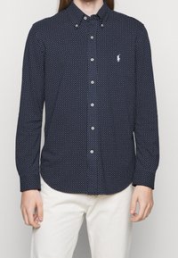 Marinblå skjorta med knappar och små vita prickar, klassisk krage och en logotyp på bröstet. Långa ärmar och en rundad nederkant.