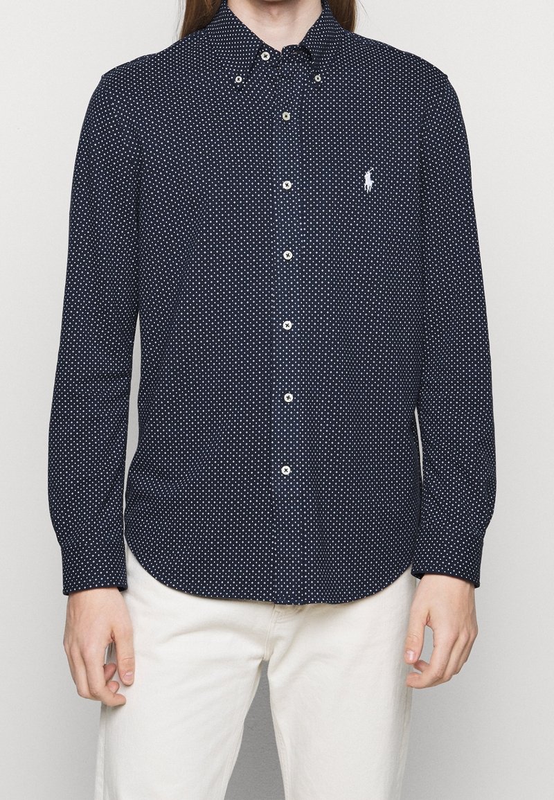 Marinblå skjorta med knappar och små vita prickar, klassisk krage och en logotyp på bröstet. Långa ärmar och en rundad nederkant.