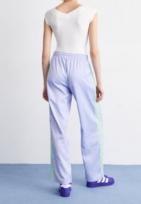 Ljuslila sportbyxor med ett mönstrat textur, elastisk midja och neongröna sidostrekar, kombinerat med lila skor med vita accenter.