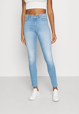 Femme portant un jean skinny taille haute bleu clair, un top court blanc et des baskets blanches, debout devant un fond uni.