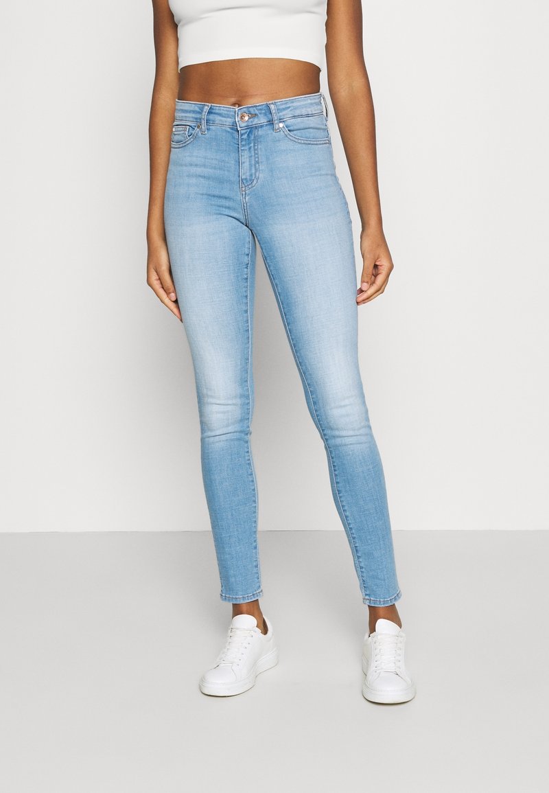 Femme portant un jean skinny taille haute bleu clair, un top court blanc et des baskets blanches, debout devant un fond uni.