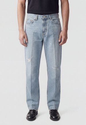 Uomo che indossa jeans dritti strappati azzurri chiari, scarpe nere e camicia nera, in piedi davanti a uno sfondo semplice.