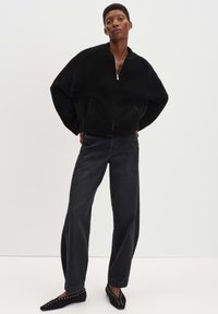 Giacca bomber nera, in tessuto strutturato, con chiusura a zip e due tasche laterali, abbinata a jeans neri a vita alta e gamba larga, insieme a décolleté nere a punta.