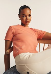 Camiseta de manga corta color coral hecha de tela suave con un acento de texto metálico en la parte inferior izquierda. Llevada con pantalones de color claro.