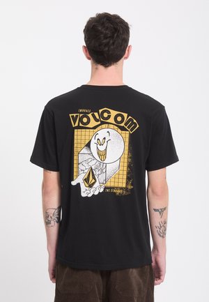 Homme portant un t-shirt noir avec un graphisme jaune et blanc d'un visage souriant et d'une main tenant une forme géométrique dans le dos.