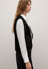 Giubbotto di velluto nero con ricami intricati, indossato sopra una camicia bianca con colletto. Capelli lunghi e mossi lasciati sciolti. Sfondo semplice.