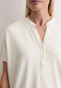 Haut à manches courtes couleur crème avec un col arrondi, une patte de boutonnage et une texture de tissu douce et lisse.