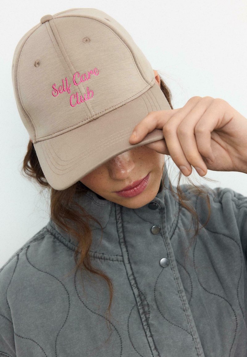 Persona con giacca grigia che inclina un berretto beige ricamato con la scritta rosa "Self Care Club", coprendosi parzialmente il volto con la mano.