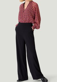 Bluse med et geometrisk mønster i pink og lilla, lange ærmer og V-hals, parret med højtaljede sorte vidde bukser.