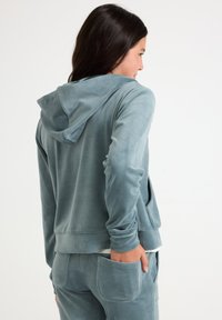 Jauns cilvēks skatās uz labo pusi, ģērbies zilonīgā velūra kapucē un atbilstošās biksēs, ar vienu roku kabatā pret baltu fona.