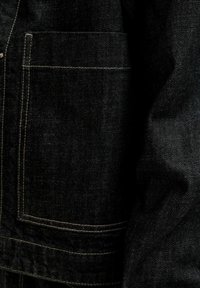 Κοντινή λήψη μαύρου τζιν υφάσματος με ορθογώνια ραμμένη τσέπη και ορατές λεπτομέρειες ραφών στο πλάι.