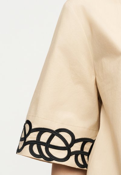 Manche courte beige avec motif de boucle noire brodée enchevêtrée le long du poignet, montrant la texture du tissu et les détails des coutures.