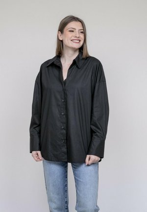 Femme souriante aux cheveux lisses mi-longs, portant une chemise noire oversize boutonnée et un jean bleu clair, debout devant un fond uni.