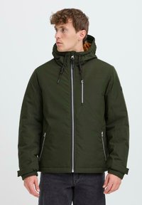 Giacca isolante verde con cappuccio, dotata di una cerniera frontale, tasche laterali con cerniera e una texture morbida. Accentuata con rivestimenti marroni.