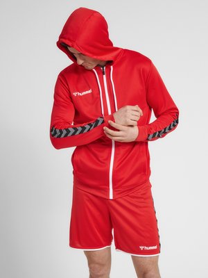 Mann trägt rote Sportjacke mit Kapuze und passende Shorts mit schwarzen Chevron-Streifen an Ärmeln und Seiten, richtet sein linkes Handgelenk.