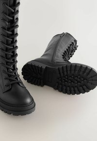 Stivali da combattimento in pelle nera presentano un design alto con lacci visibili, una cerniera laterale e una suola in gomma resistente e testurizzata con scanalature profonde.