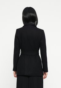 Mujer con el pelo lacio y negro, vistiendo un blazer negro ajustado con cinturón y una falda negra con pliegues, de pie contra un fondo blanco.
