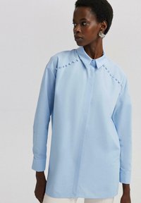 Camisa de botones de color azul claro con un collar estructurado, mangas largas y detalles decorativos de pompones a lo largo de la costura del hombro. Tejido suave.