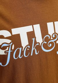 Brązowa koszulka bawełniana z dużymi białymi literami i niebieskimi akcentami w stylu pisma ręcznego, z napisem "STU Jack &". Miękka w dotyku, swobodny design.