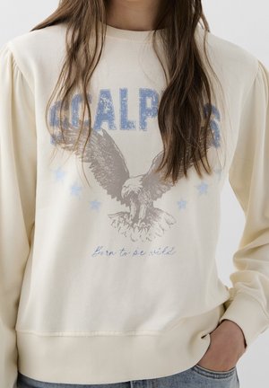 Femme portant un sweat-shirt crème avec un texte bleu délavé « SCALPERS », un graphisme d'aigle et l'inscription « Born to be wild », main dans la poche, cheveux longs bruns.