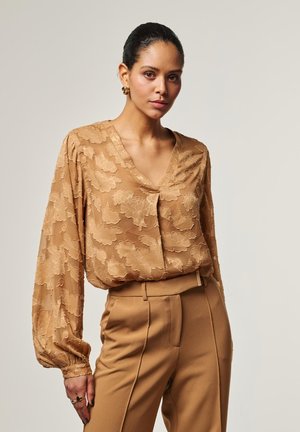 Femme portant un chemisier à manches longues texturé à motifs floraux beige, rentré dans un pantalon taille haute beige, posant devant un fond uni.