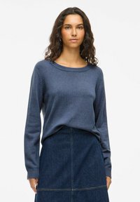 Temno modra pletena pulover s okroglo vratno otvoritvijo in dolgimi rokavi, nošen preko jeansa krila. Gladka tekstura in sproščen kroj.