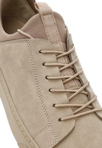 Sneaker beige in suede con superficie testurizzata, lacci piatti e un'etichetta sul tallone intrecciata. Presenta cuciture a vista e suola in gomma.