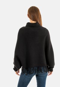 Pull en tricot surdimensionné noir avec une texture côtelée, un col haut et des franges au bas. Associé à un jean sombre.